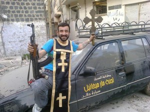 A rebel in the old city of Homs, where 80,000 Christians were forced out. He poses with a machine gun in one hand and a processional cross in the other; he is wearing part of the funeral vestments of a Syriac Orthodox priest. In this image he is seen leaning against the funeral hearse of St. Joseph’s Syriac Orthodox Parish. It is said that the rebels in the area used the hearse to transport weapons around the district.